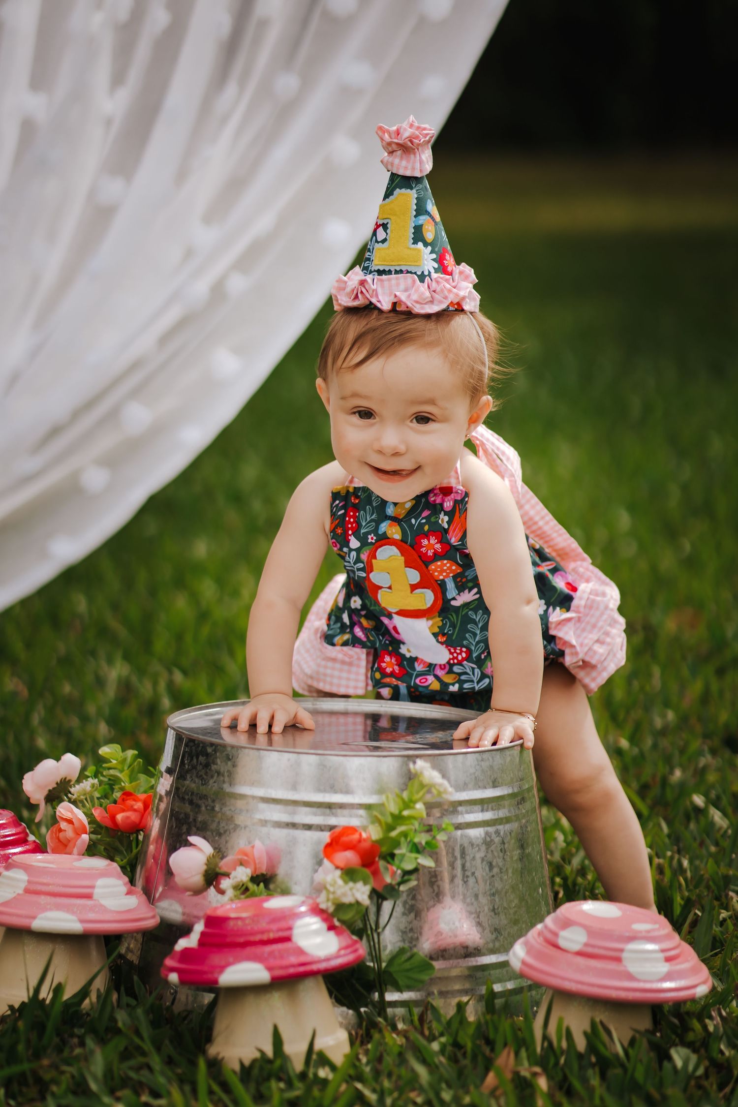 Baby girl in floral outfit and party hat smiles beside a tin bucket with decorative mushrooms on green grass.