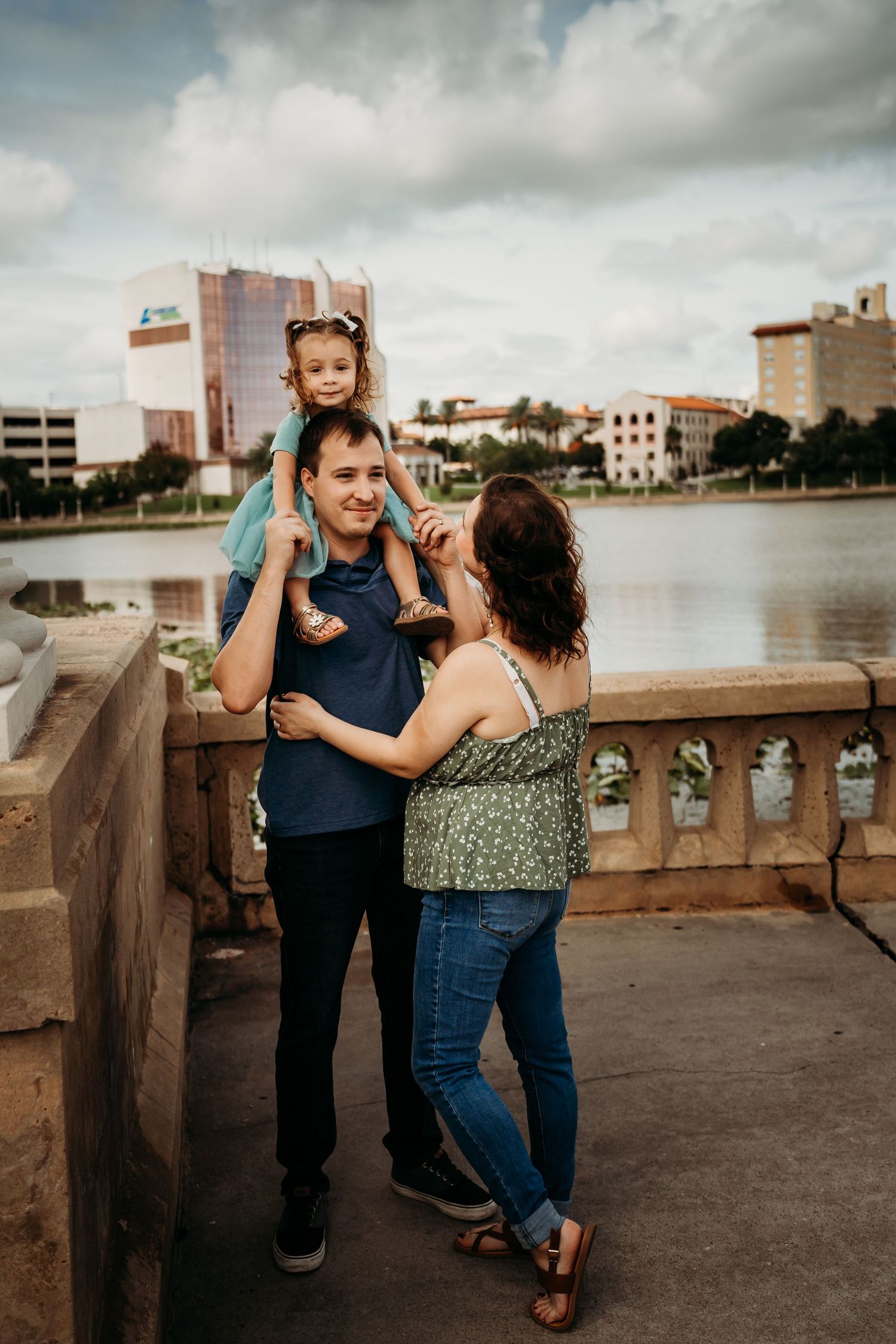 Family of three posing by a waterfront, with daughter sitting on fathers shoulders.
