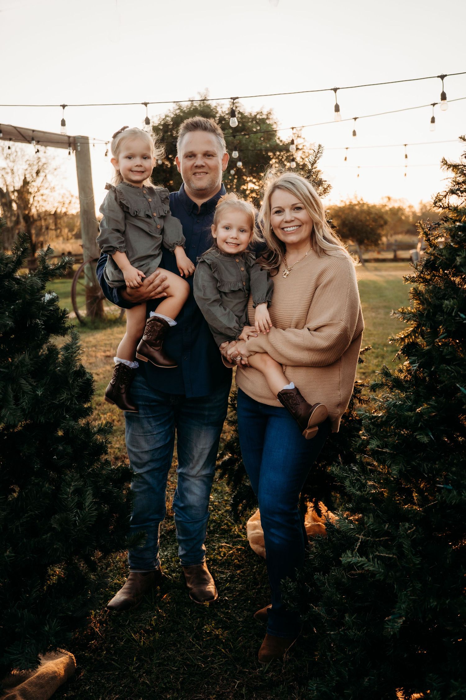 Family of four poses among Christmas trees at golden hour, parents holding toddlers in coordinating outfits.
