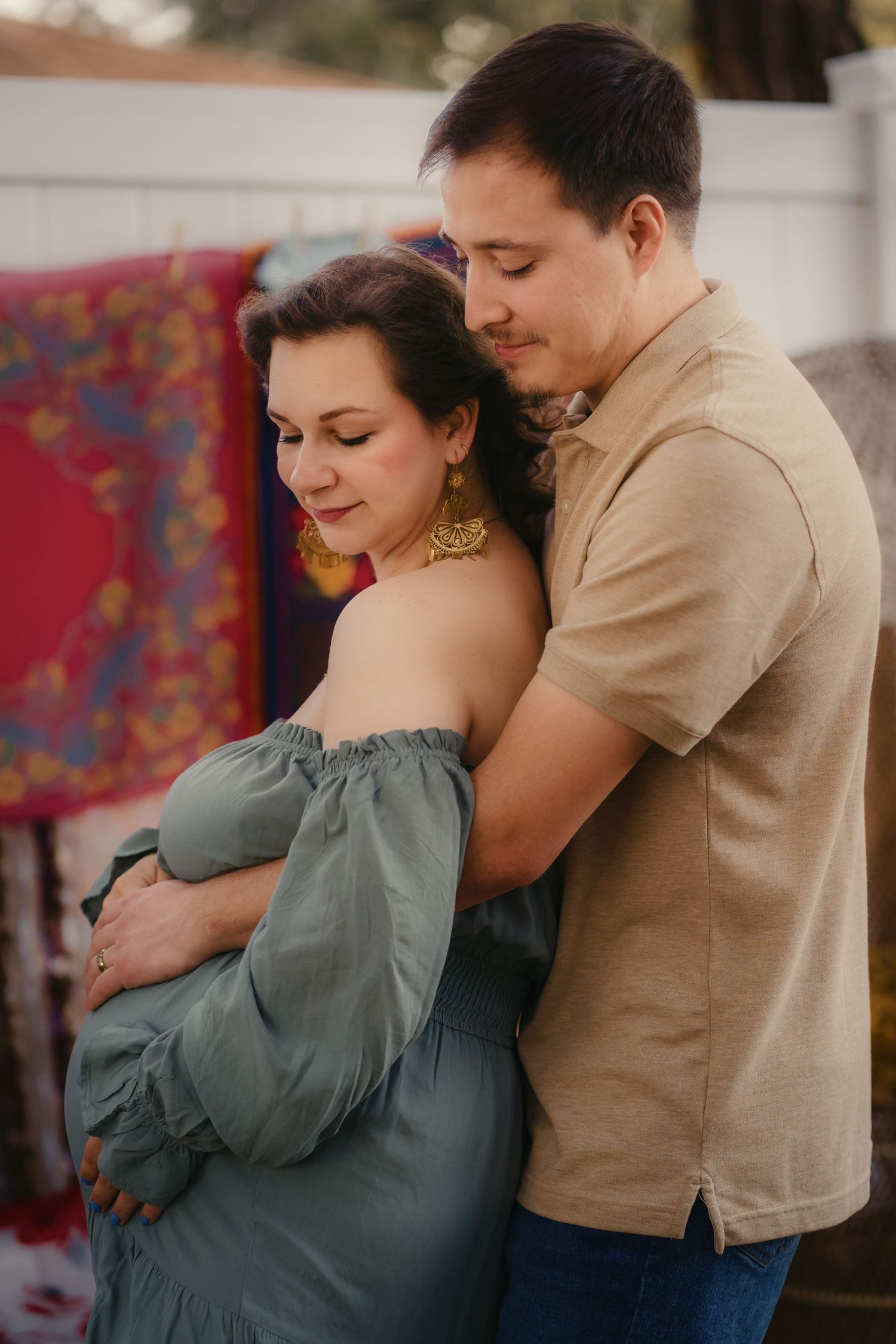 A couple shares a tender embrace outdoors, the man gently holding the pregnant woman in a teal off-shoulder dress.