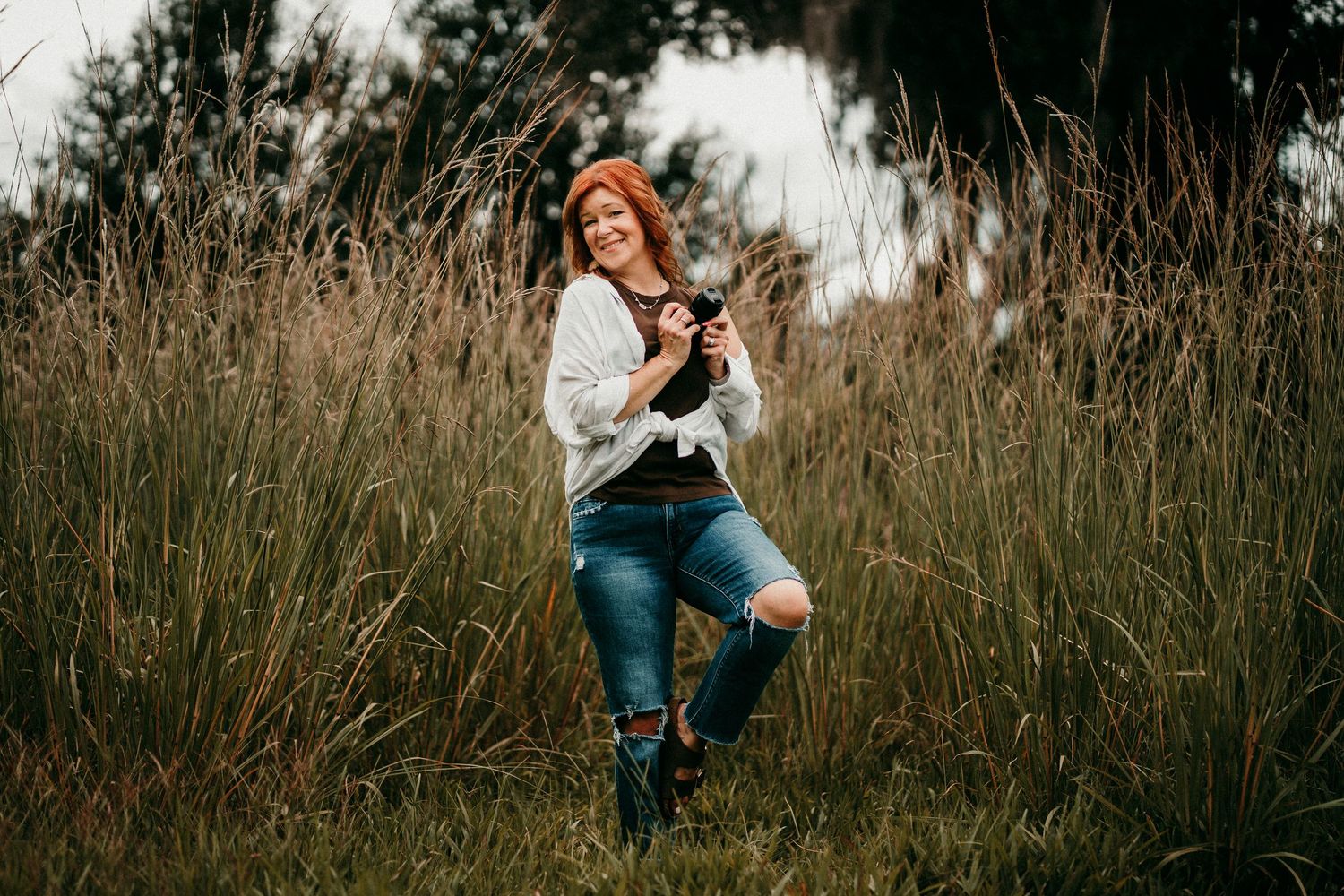 Smiling red-haired woman holding camera while standing in tall golden grass outdoors.