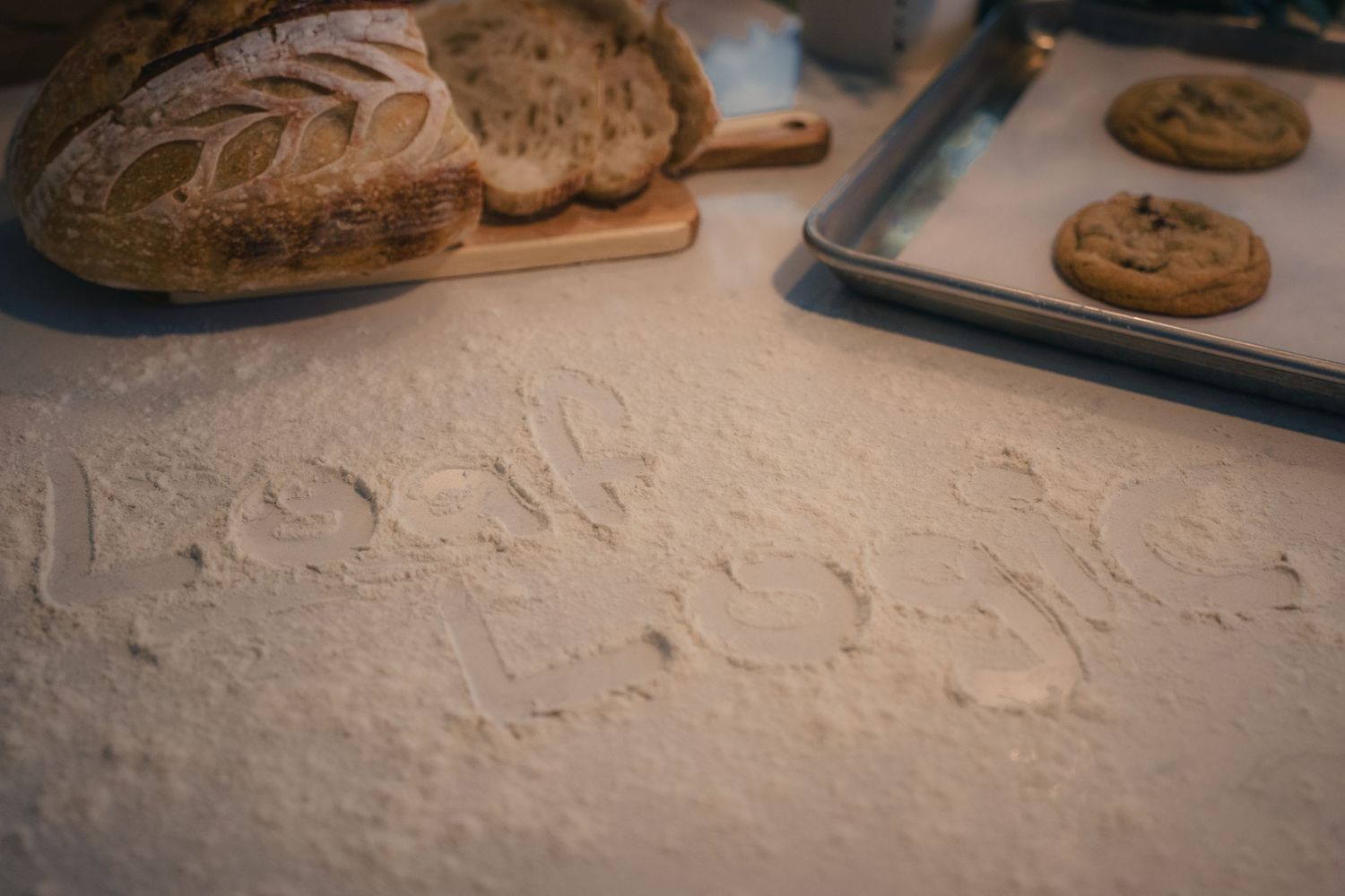 Flour dusted countertop with handwritten text, rustic sourdough bread on cutting board, and chocolate chip cookies on baking sheet.