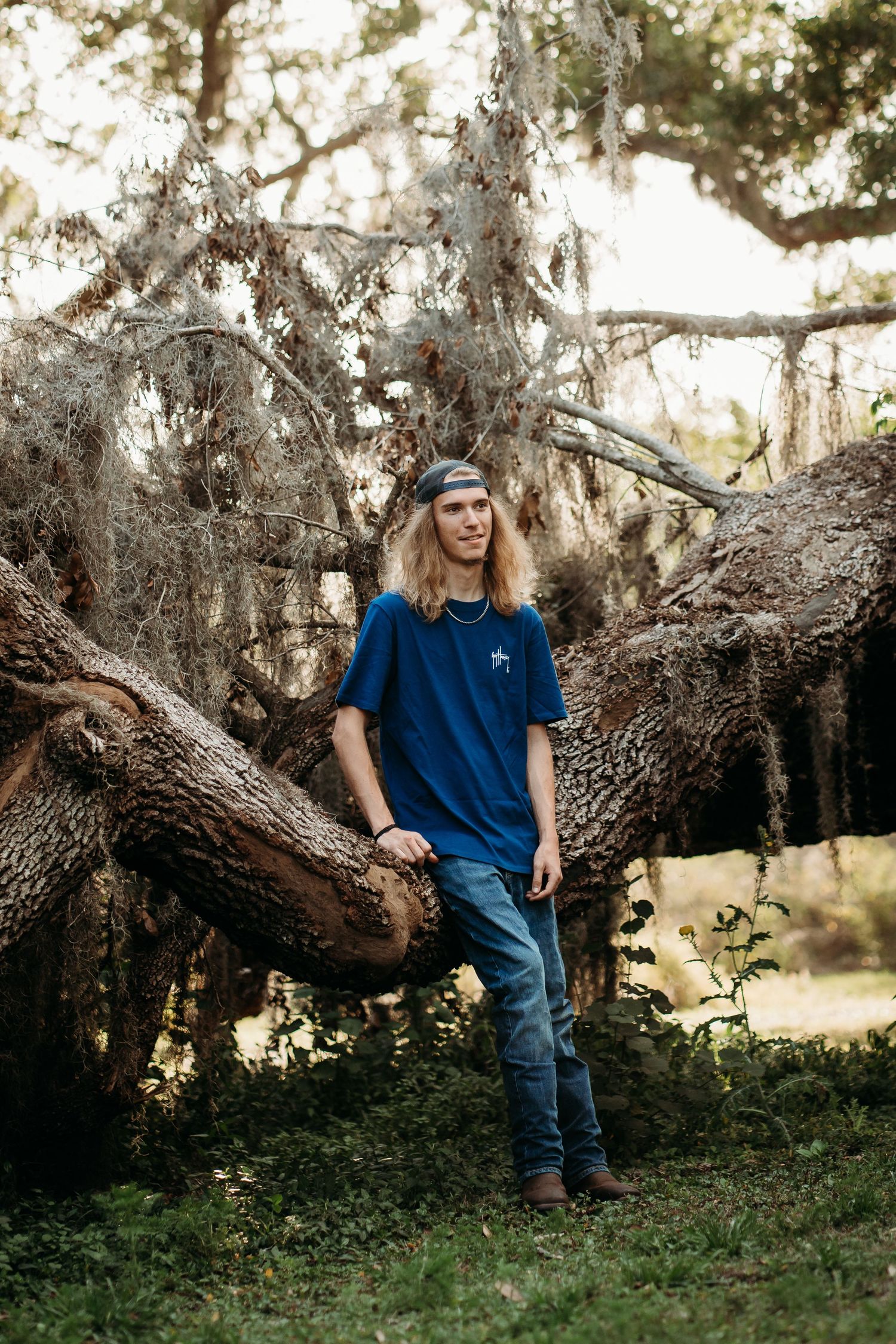 Young man with long blonde hair in blue t-shirt and jeans leaning against a large moss-covered oak tree outdoors.