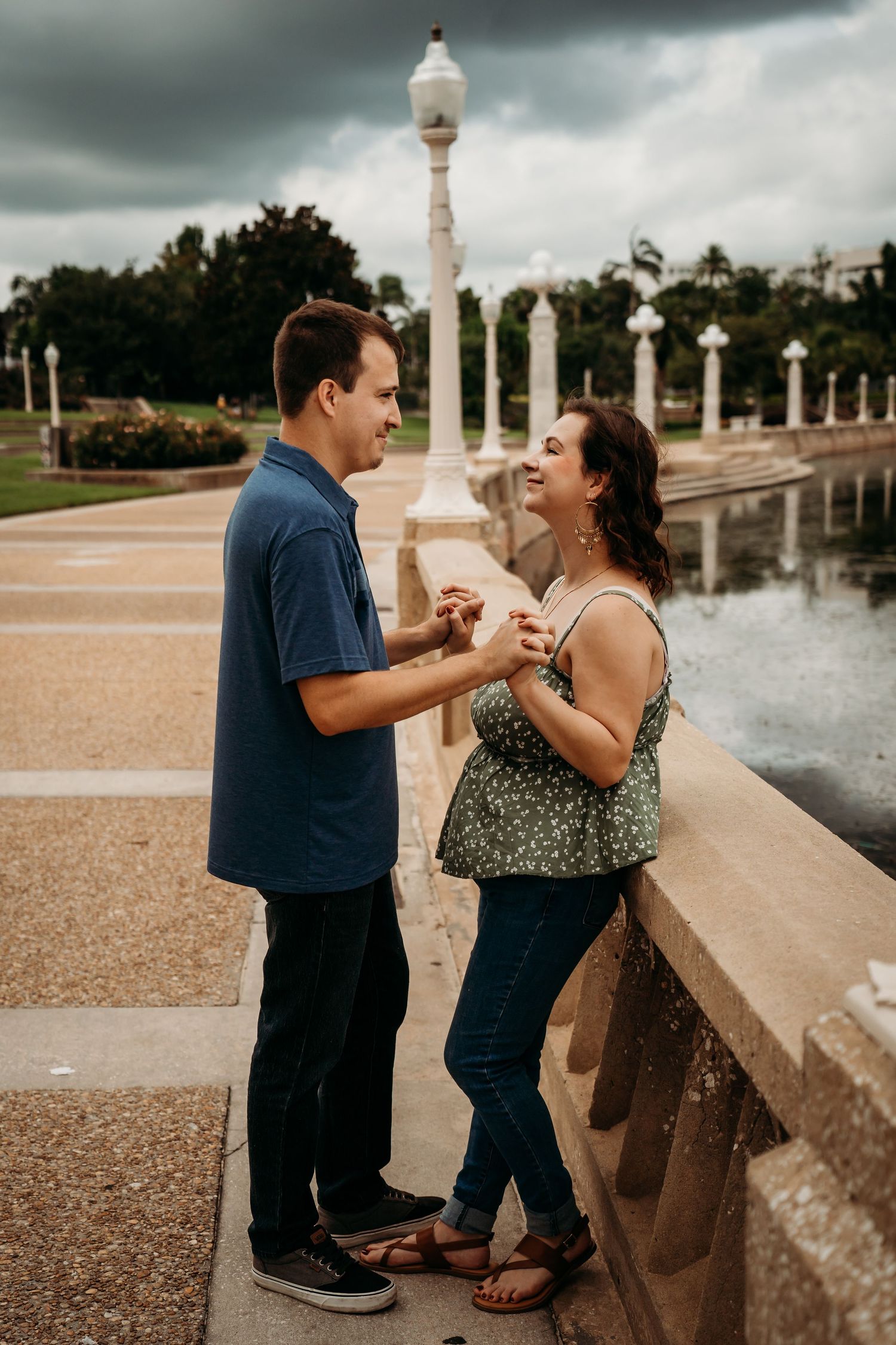 Couple holding hands and smiling at each other on a stone bridge in a park on a cloudy day.