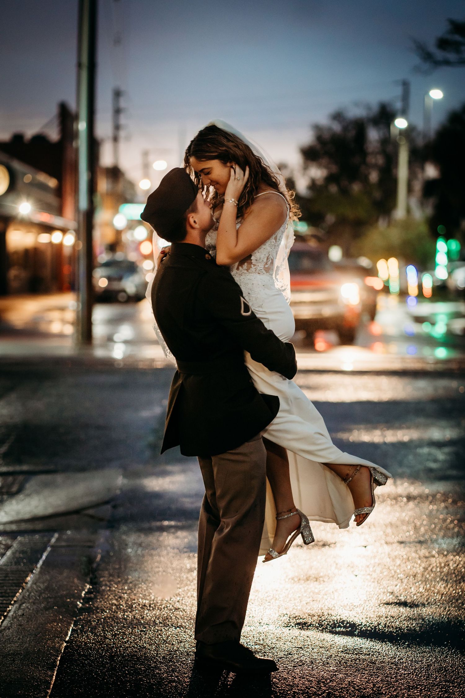 Groom lifting bride in wedding attire on a rain-slicked city street at night with bokeh lights.