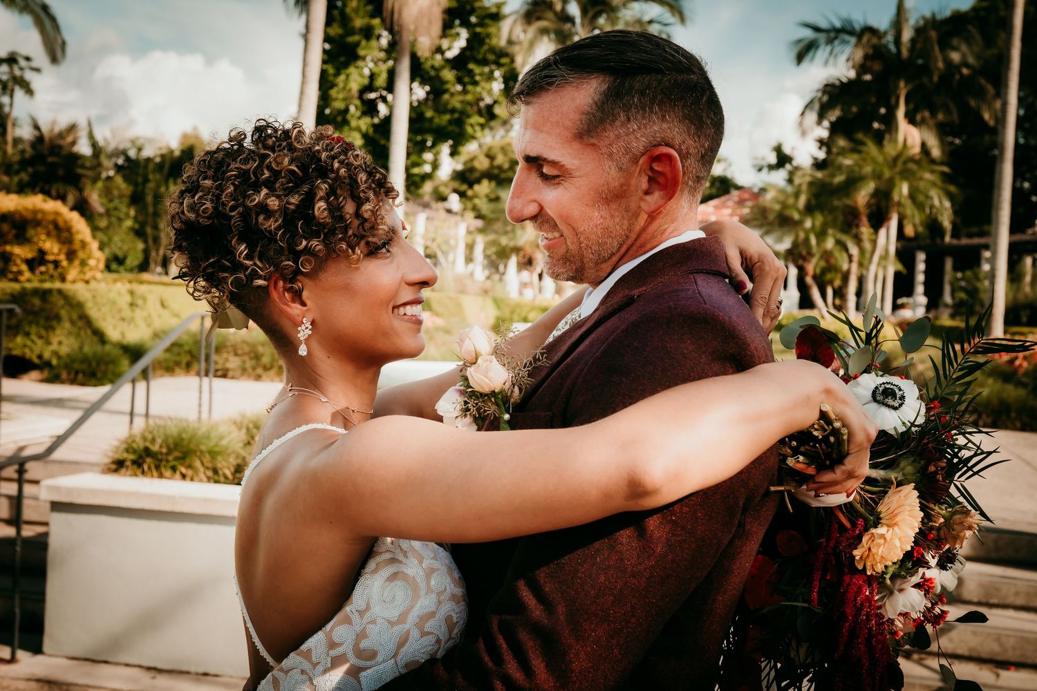 A joyful newlywed couple embraces outdoors, bride in lace gown with curly hair, groom in burgundy suit, tropical setting.