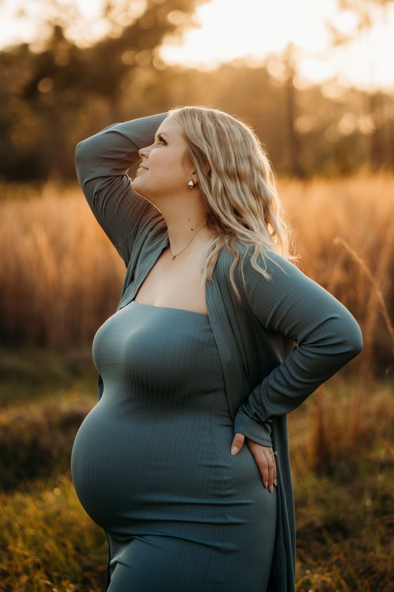 Pregnant blonde woman in teal dress poses gracefully in golden field at sunset.