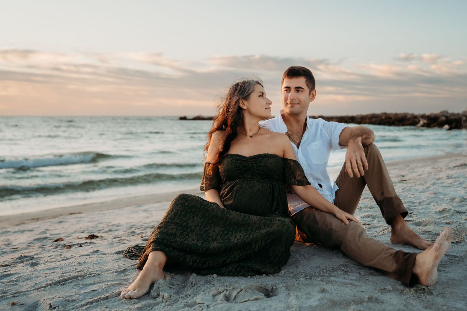 A pregnant couple sits together on a sandy beach at sunset, the woman in a dark off-shoulder maternity gown.
