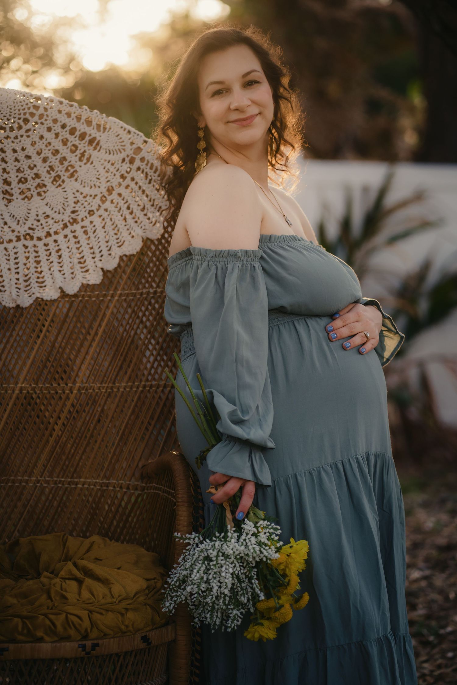 Pregnant woman in off-shoulder sage dress holding wildflowers, posing by wicker chair at golden hour.