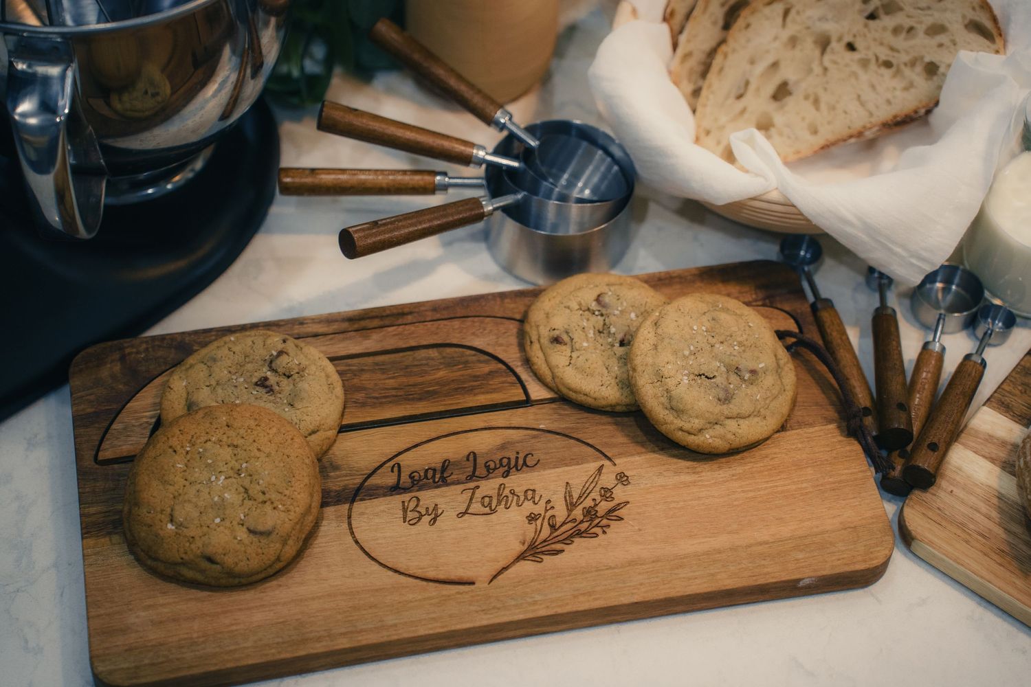 Personalized wooden cutting board engraved with 'Just Logic By Lohn' displayed with chocolate chip cookies and cinnamon sticks.