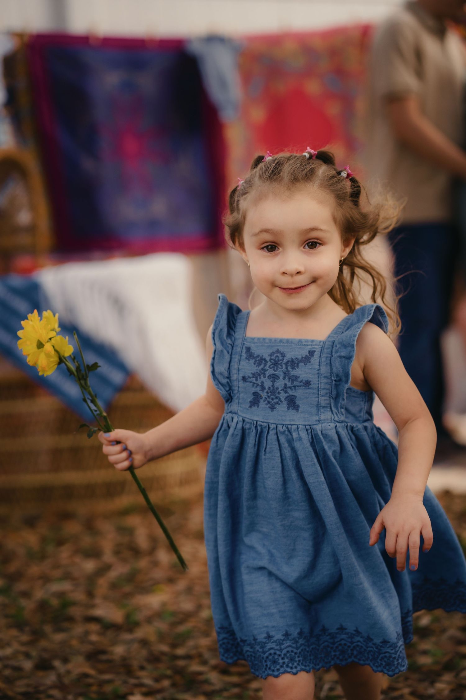 A smiling little girl in a blue embroidered dress holds yellow flowers at a colorful outdoor market.