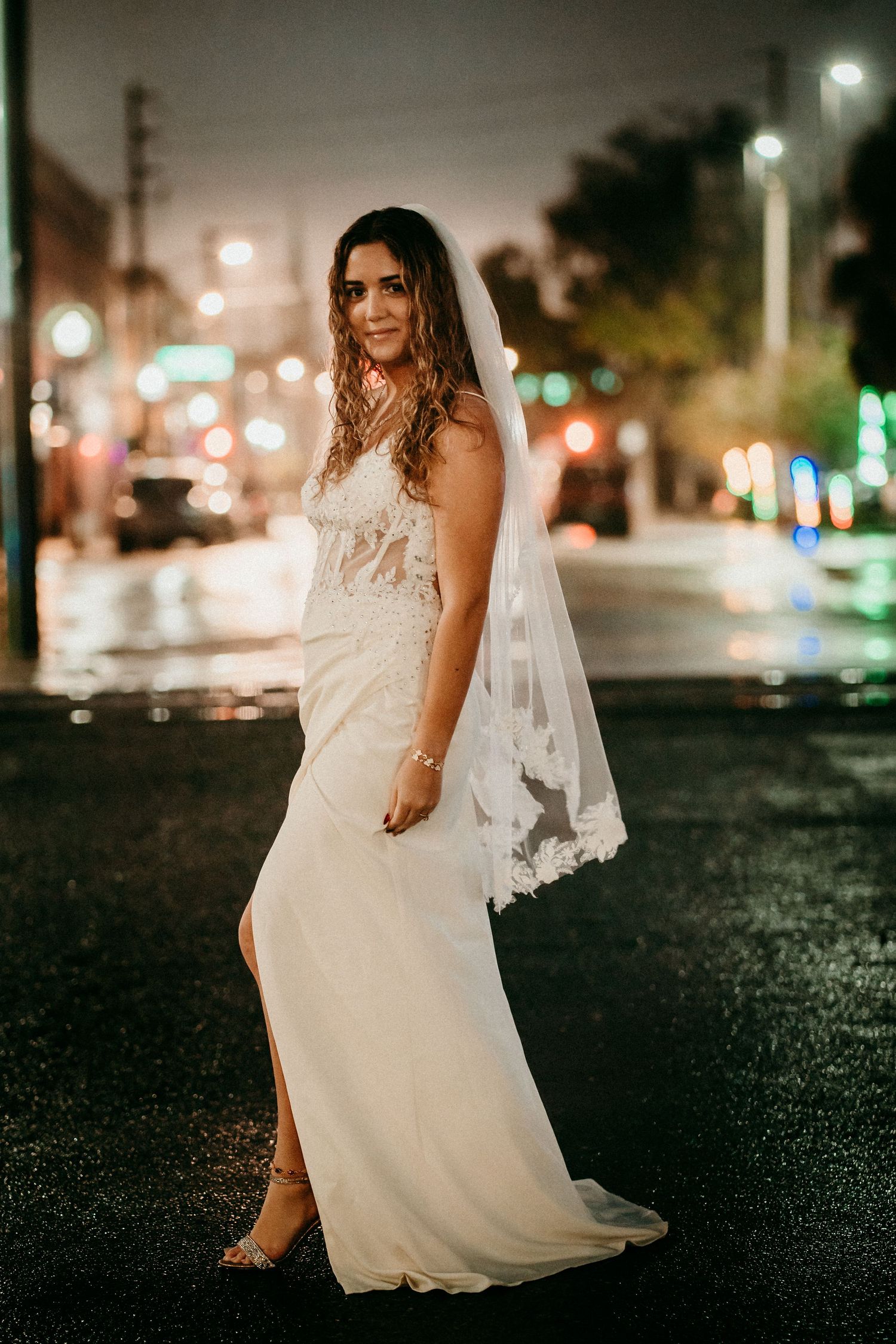 Bride in white lace gown and veil poses on wet city street at night with bokeh lights.