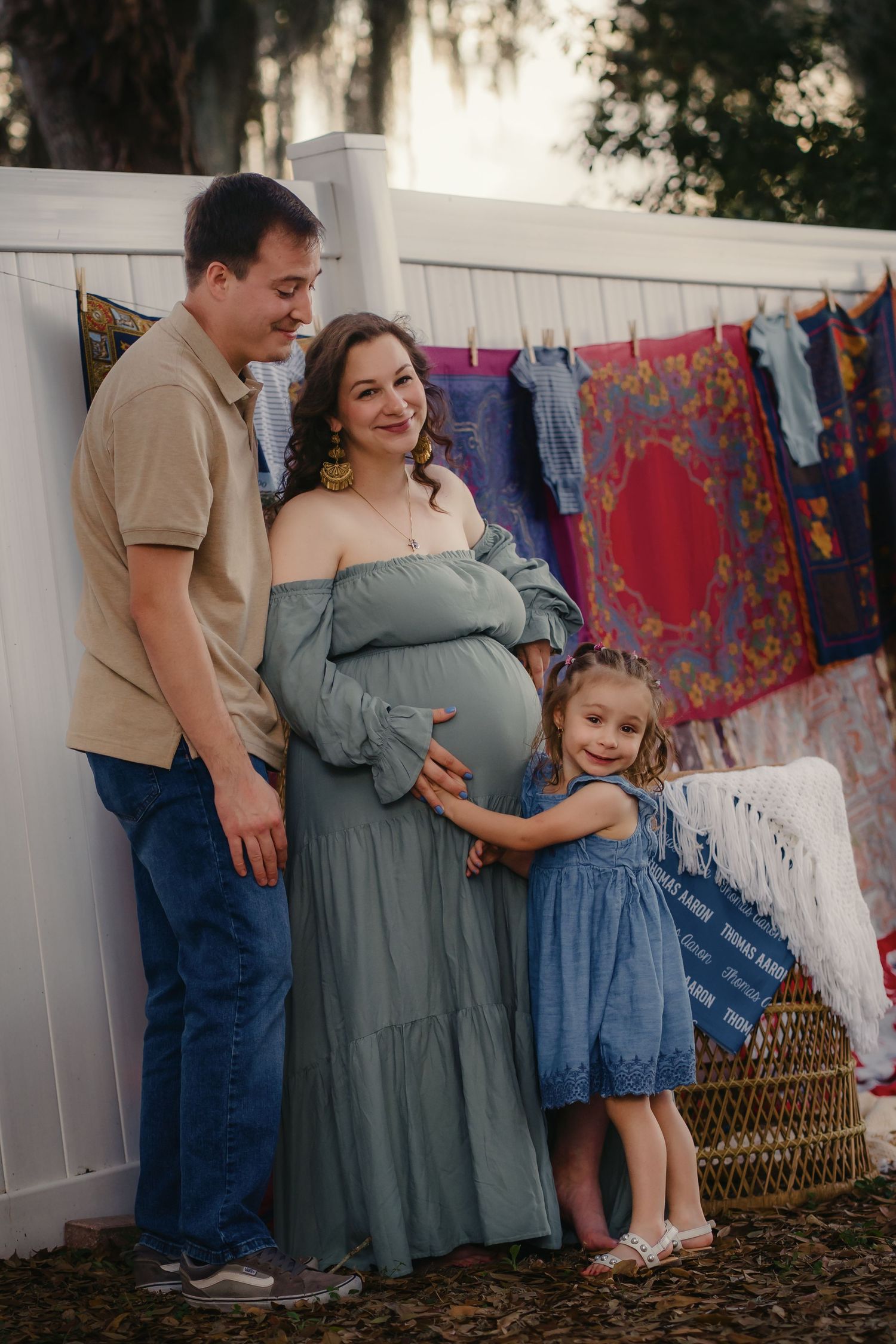 A pregnant woman in a teal dress poses with her husband and young daughter during a maternity photo session outdoors.