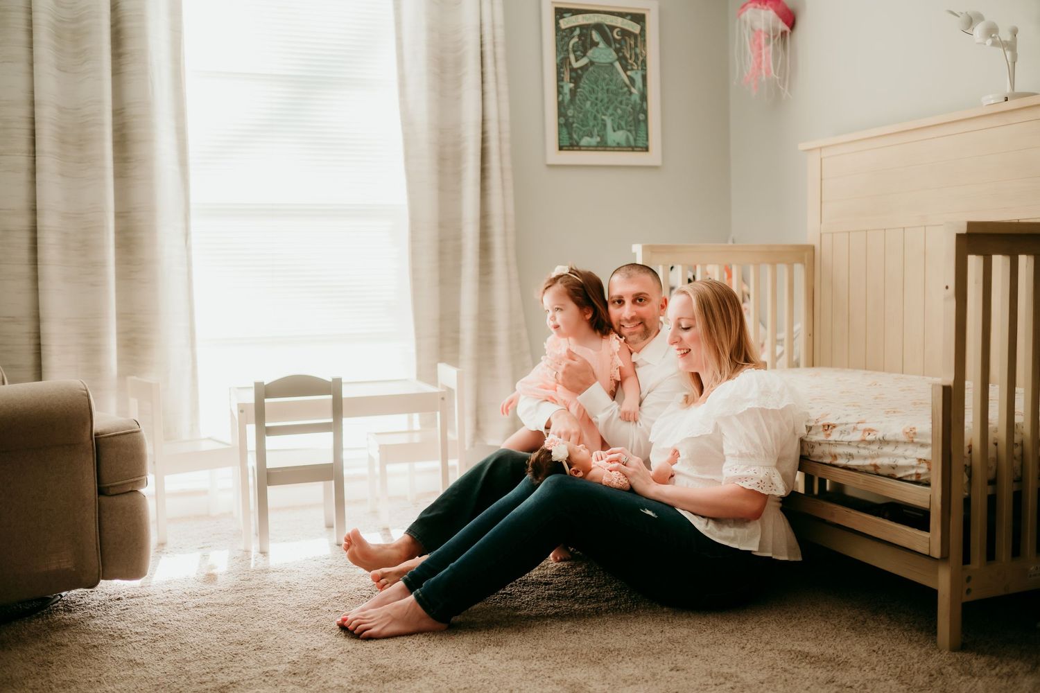 A happy family sits on a nursery floor, parents smiling with their toddler daughter in a warmly lit room.