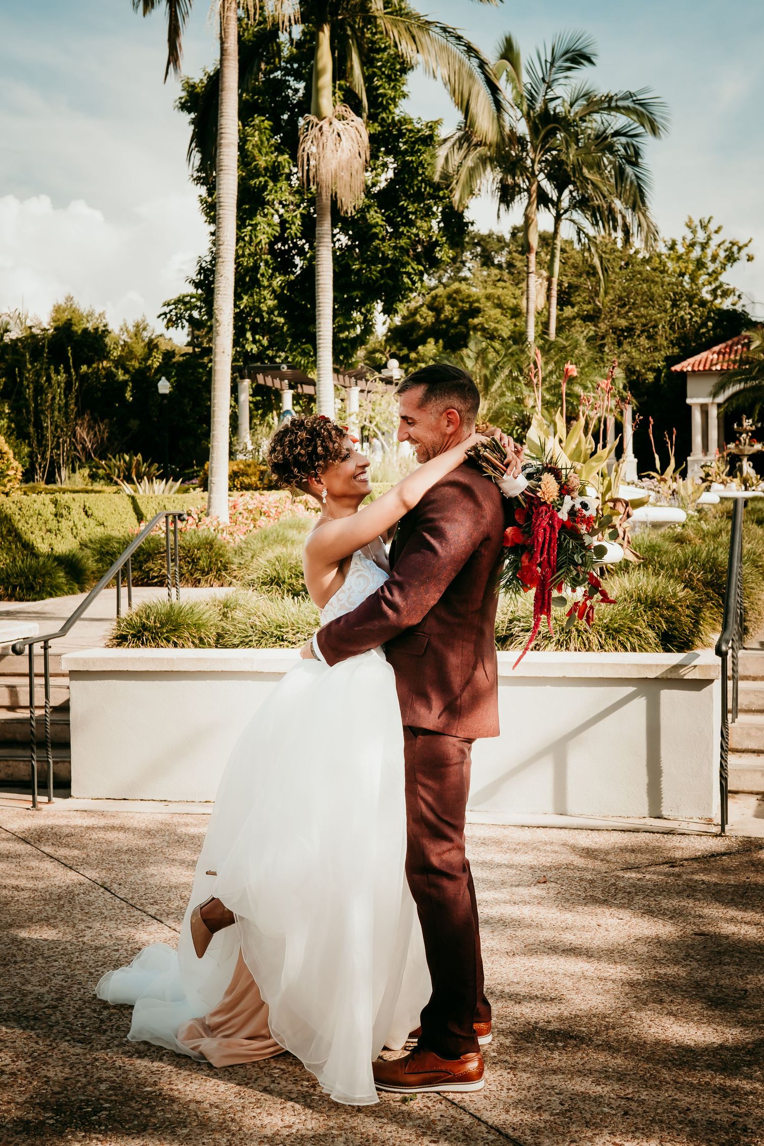Bride and groom embrace outdoors at tropical wedding venue, she holds bouquet as he lifts her in maroon suit.