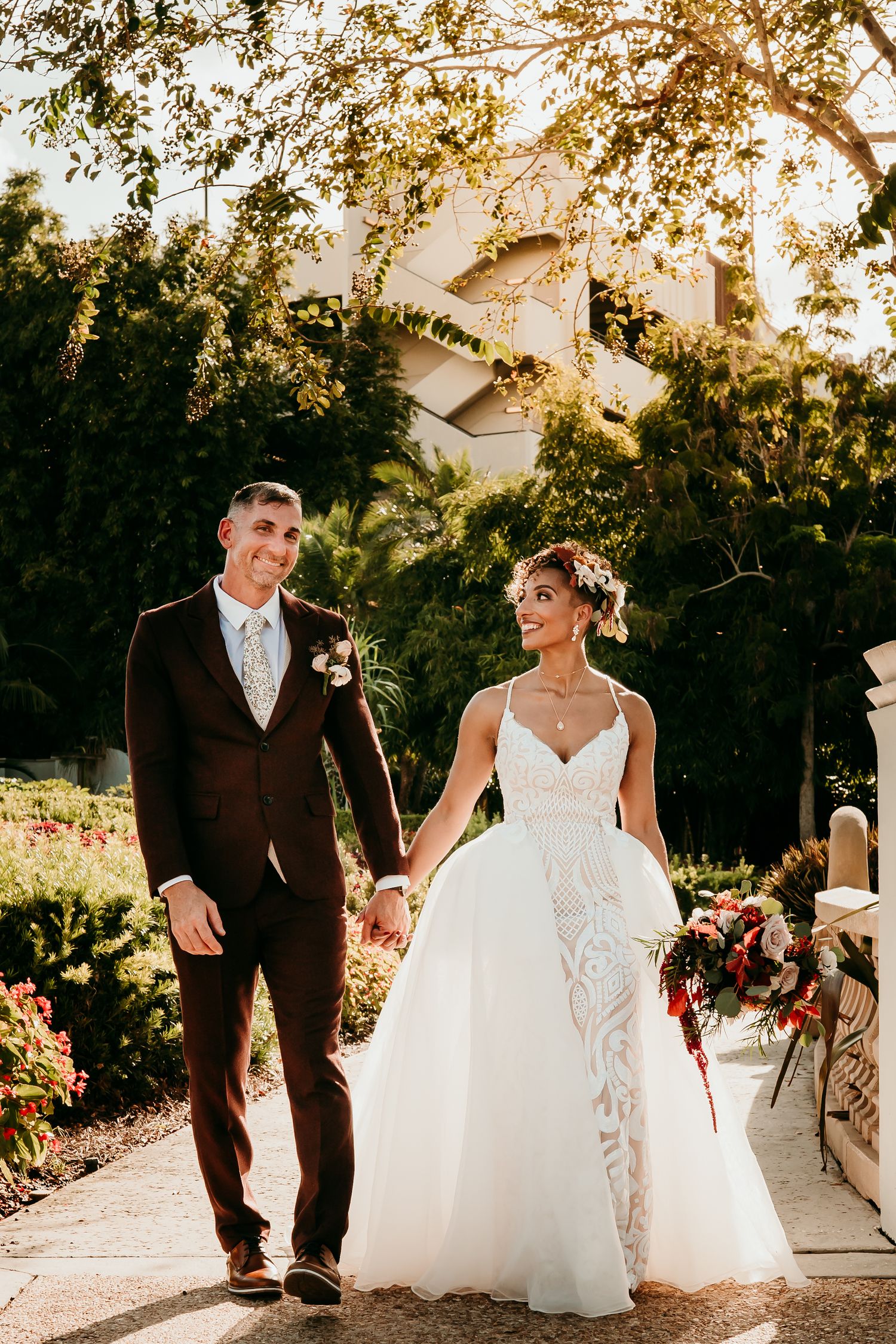 Bride and groom walk hand-in-hand through a sunlit garden, she in a lace ballgown, he in a dark suit.