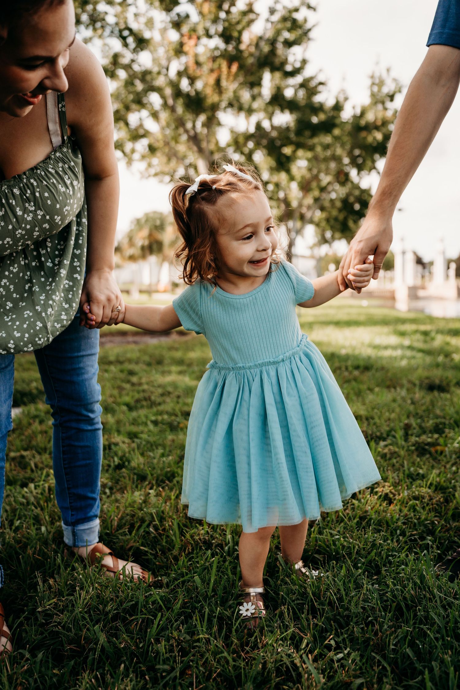 Smiling toddler girl in teal dress holding parents hands while walking on green grass.