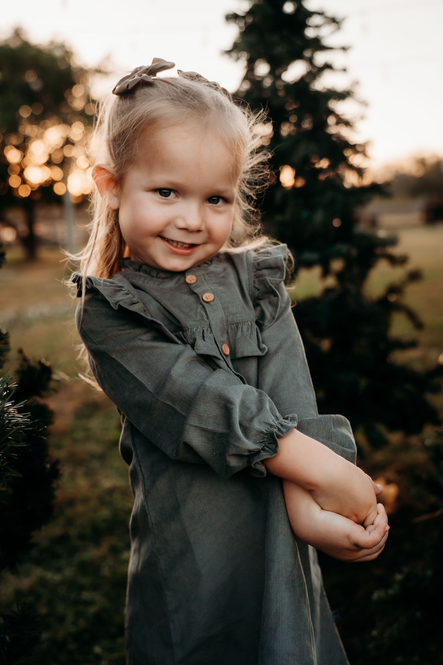 Adorable little blonde girl in gray ruffled dress smiling warmly at sunset near evergreen trees.