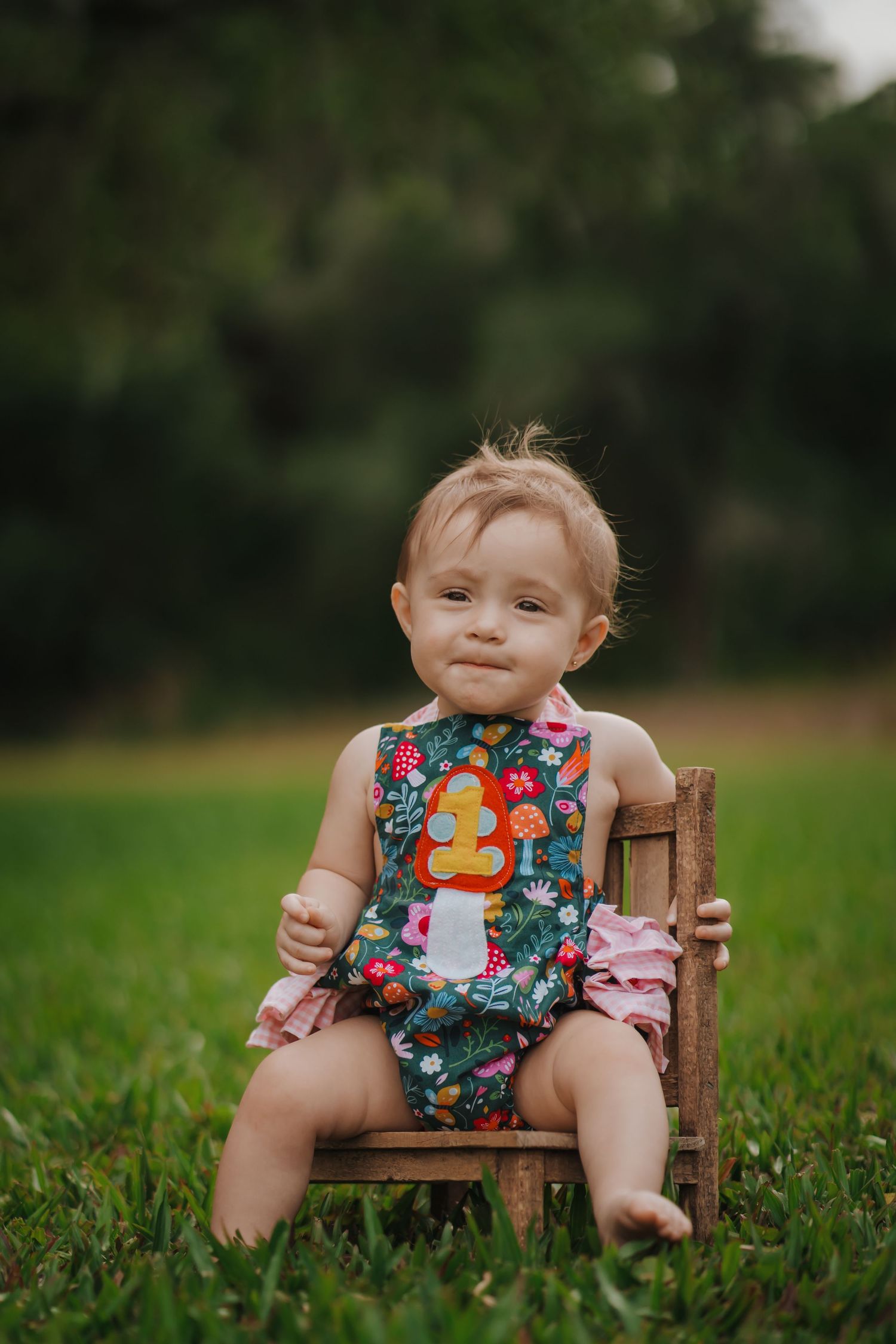 Adorable baby girl in colorful mushroom romper sitting on tiny wooden chair on green grass outdoors.