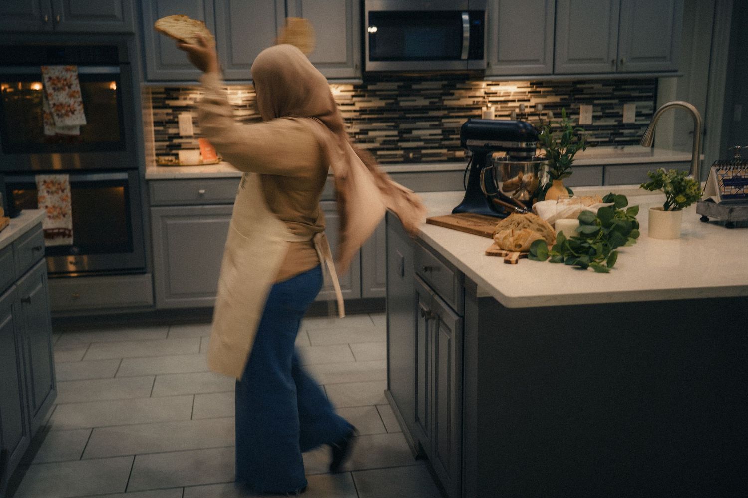 Woman tossing pizza dough in modern kitchen with gray cabinets and KitchenAid mixer on counter.