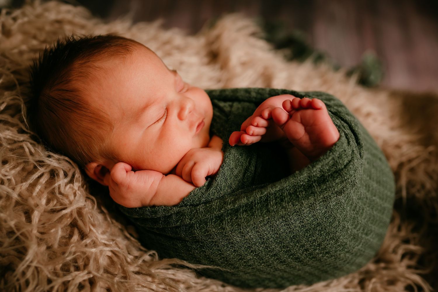Peaceful newborn baby wrapped in green knit blanket sleeping on fluffy fur backdrop.