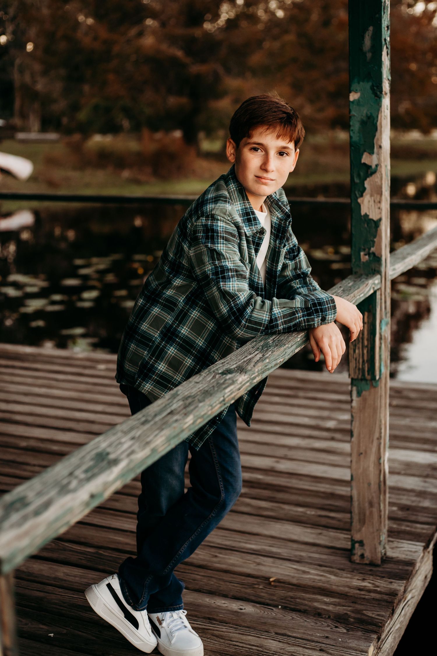 Teen boy in plaid shirt leaning on wooden dock railing by a pond in autumn park setting.