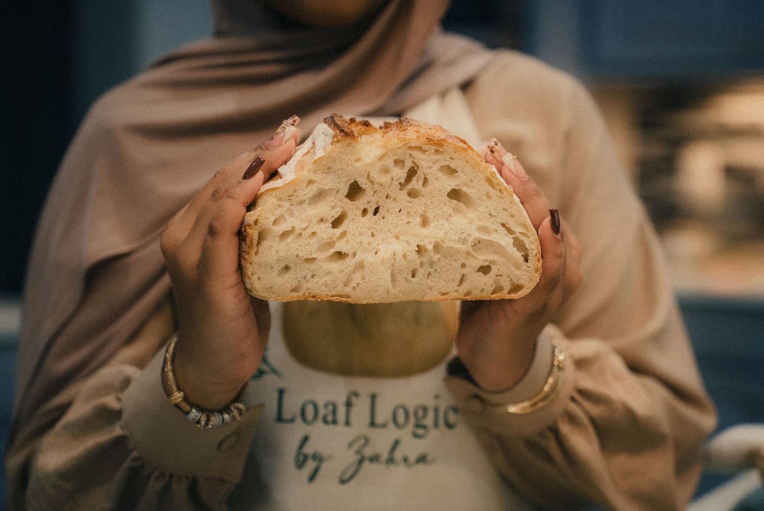 Woman in hijab holds freshly baked sourdough bread loaf, wearing a Loaf Logic by Zahra apron.