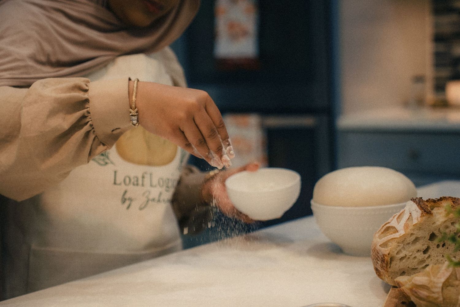 A baker in a hijab and apron dusts flour over dough on a counter at Loaf Lounge by Jaha bakery.