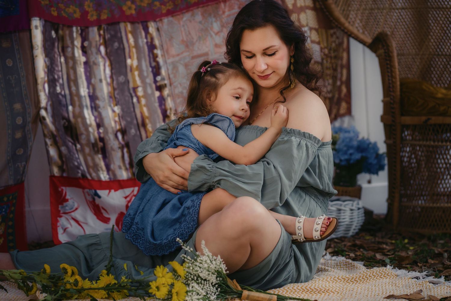 A mother tenderly embraces her young daughter in a cozy boho-styled setting with flowers and colorful textiles.