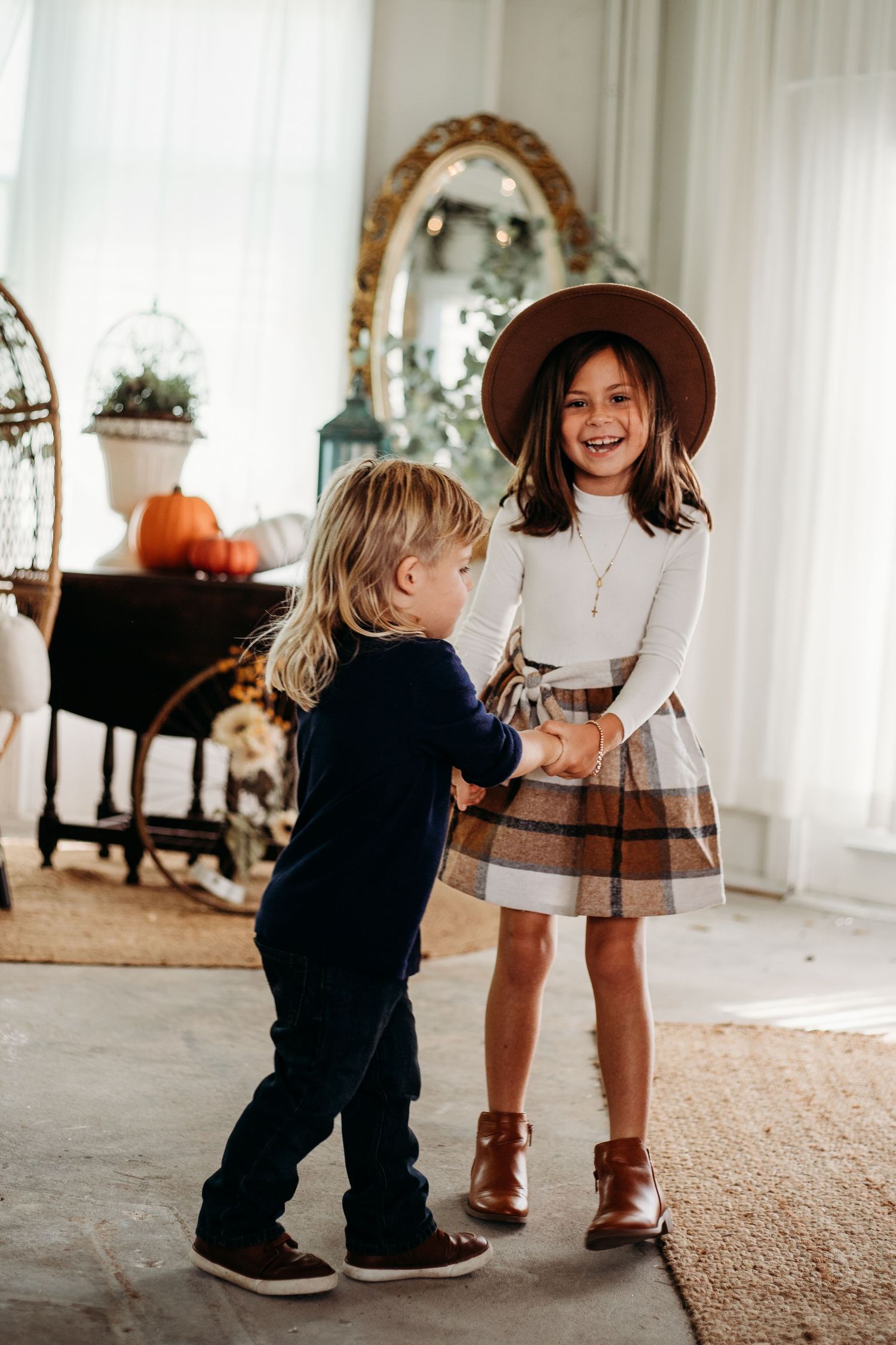 Two young children laughing and holding hands indoors, girl in plaid skirt and wide-brim hat.