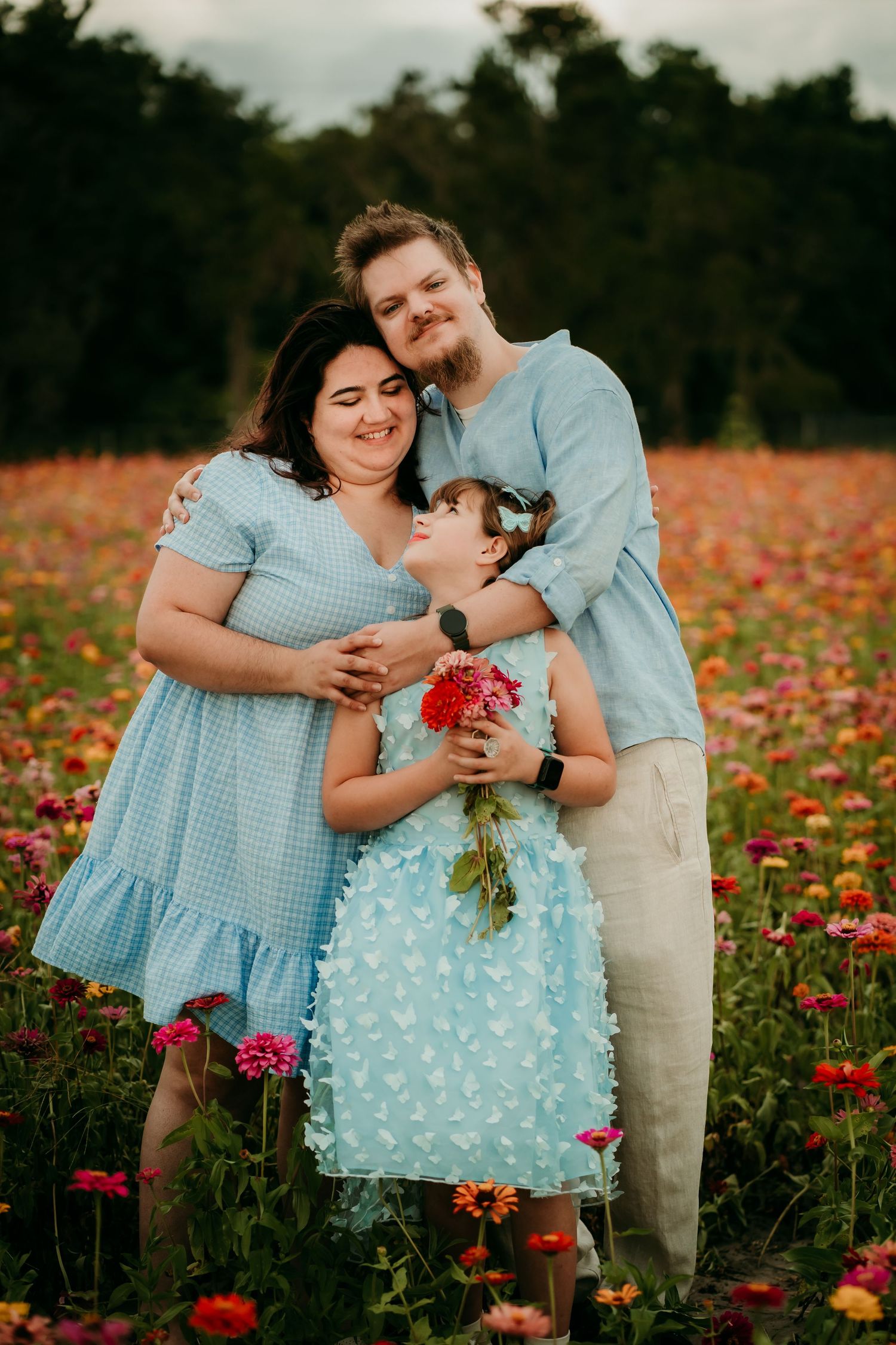 Family of three posing in a colorful flower field, wearing matching blue outfits, with a young girl holding red flowers.