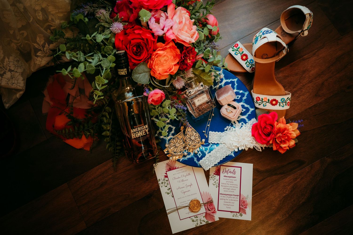 Colorful wedding flat lay with vibrant floral bouquet, embroidered heels, invitations, and whiskey bottle on dark wood floor.