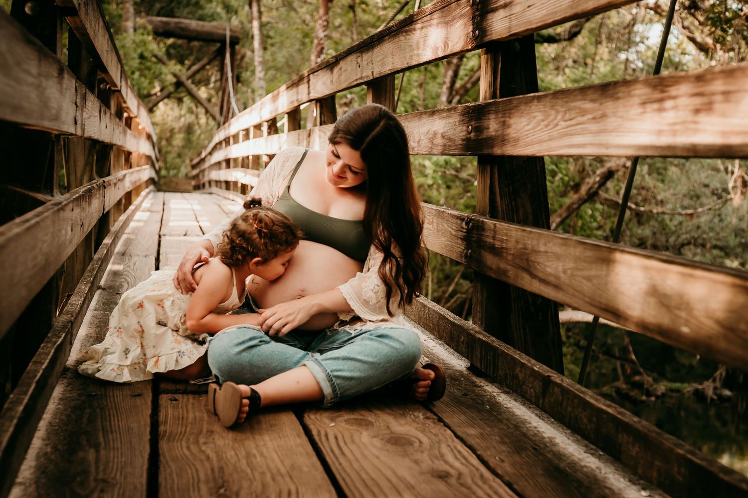 Pregnant mother sitting on wooden bridge laughing with her young toddler child outdoors.
