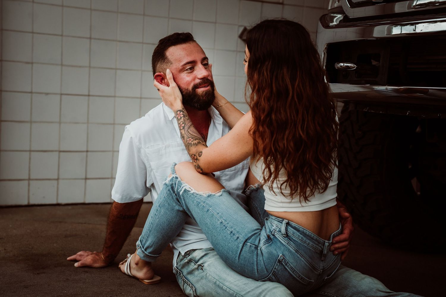 Tattooed couple sitting on garage floor, woman tenderly touching mans face in romantic moment.