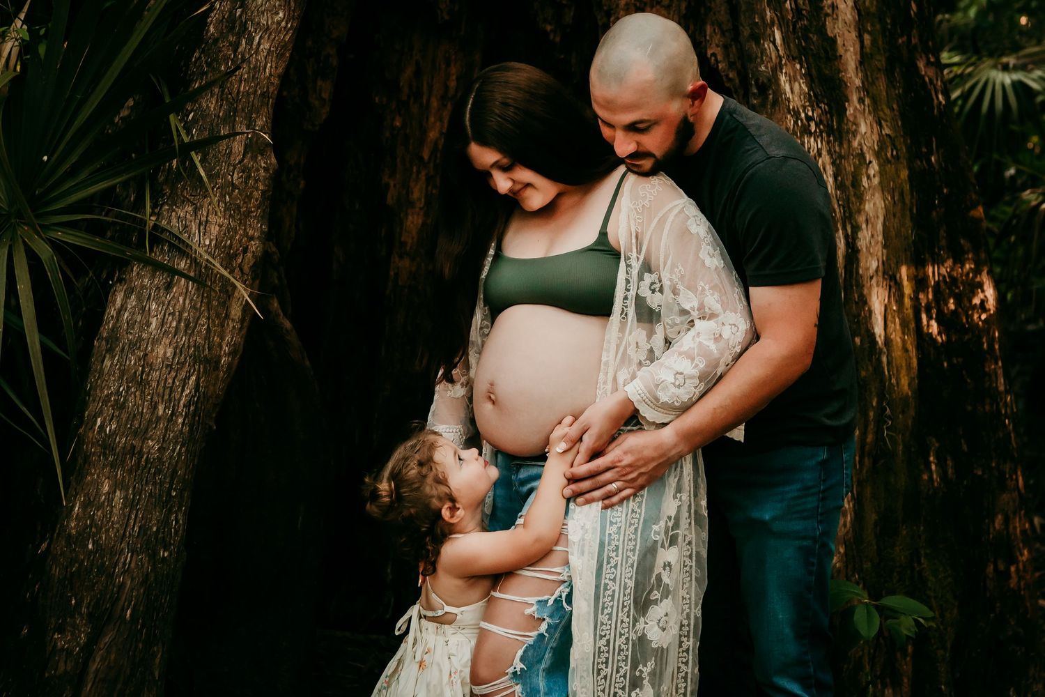 Expecting couple with toddler daughter posing in front of large tree during maternity photoshoot.
