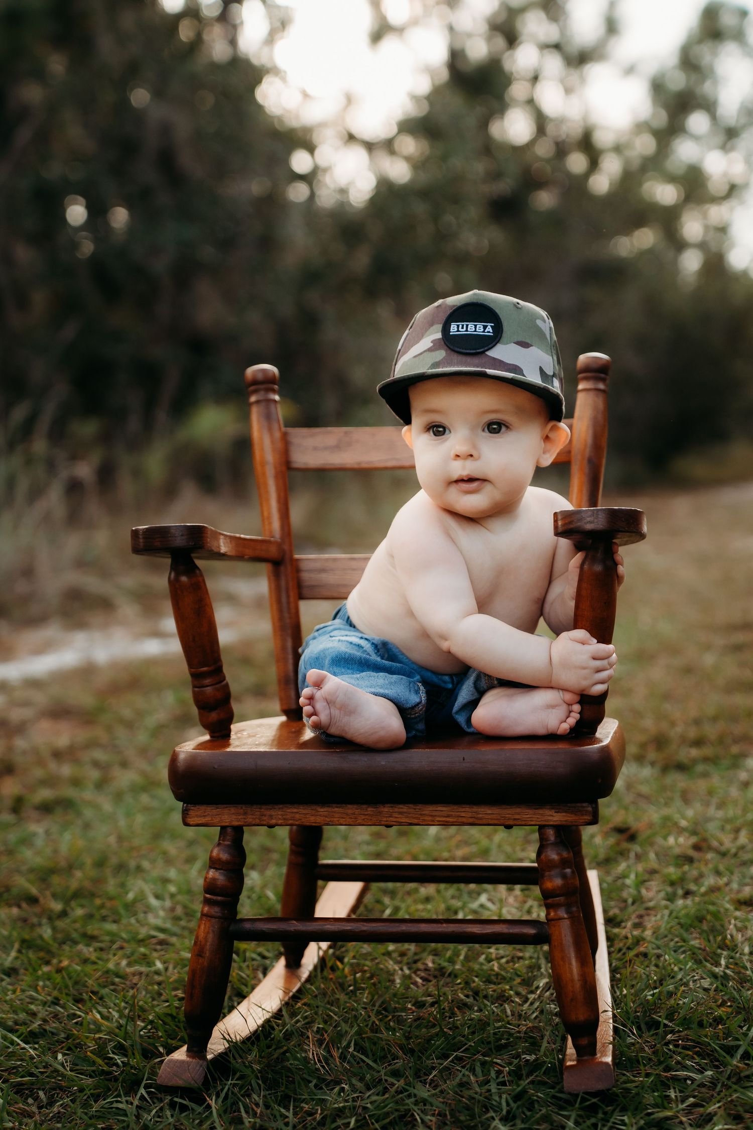 Adorable baby boy in denim shorts and camo hat sitting on a wooden rocking chair outdoors on green grass.