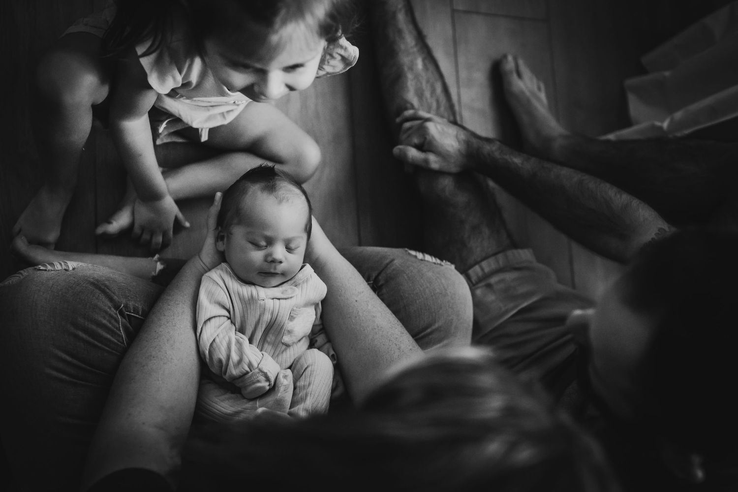 Black and white photo of a newborn baby surrounded by siblings in a warm, intimate family moment.