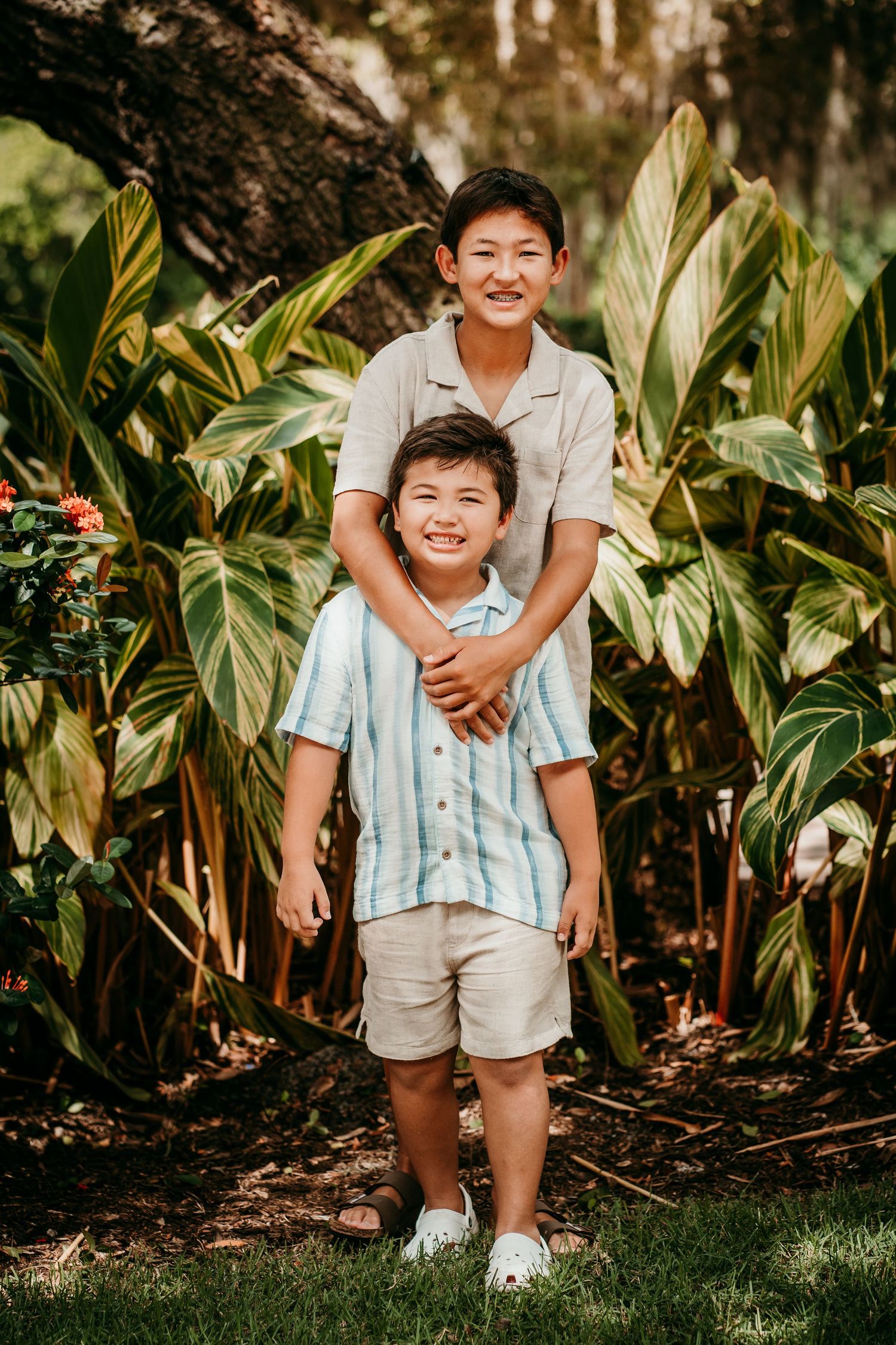 Two brothers smiling and hugging in front of lush tropical green plants outdoors.