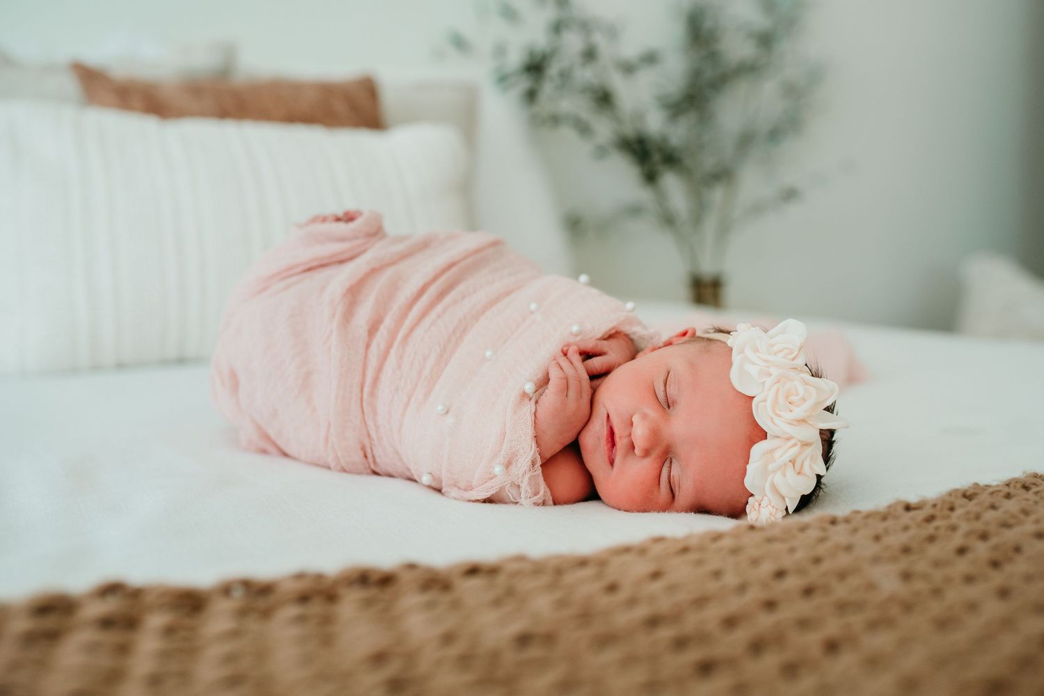 Newborn baby girl swaddled in pink wrap with white floral headband sleeping on white bed.