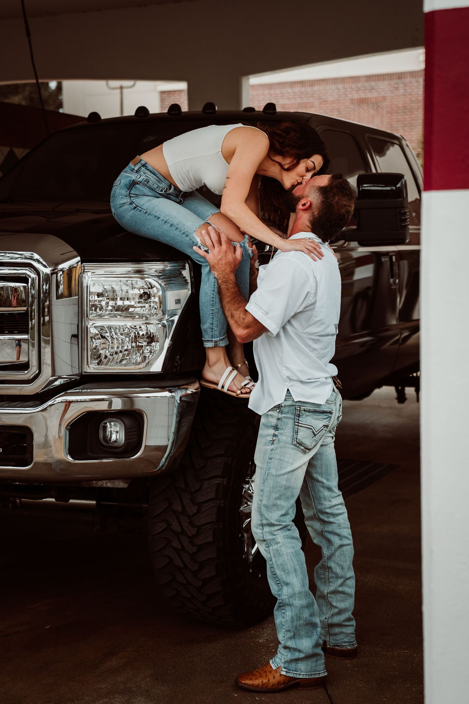 A woman sitting on a truck bumper leans down to kiss a man in a parking garage in a playful pose.