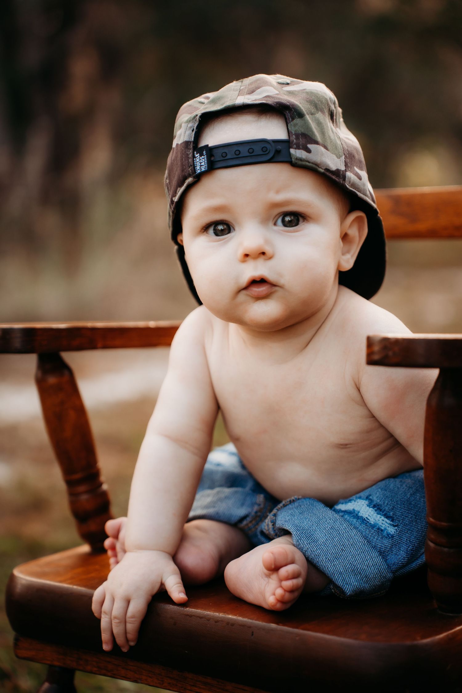 Adorable shirtless baby wearing camo snapback hat and denim shorts sitting on wooden chair outdoors.