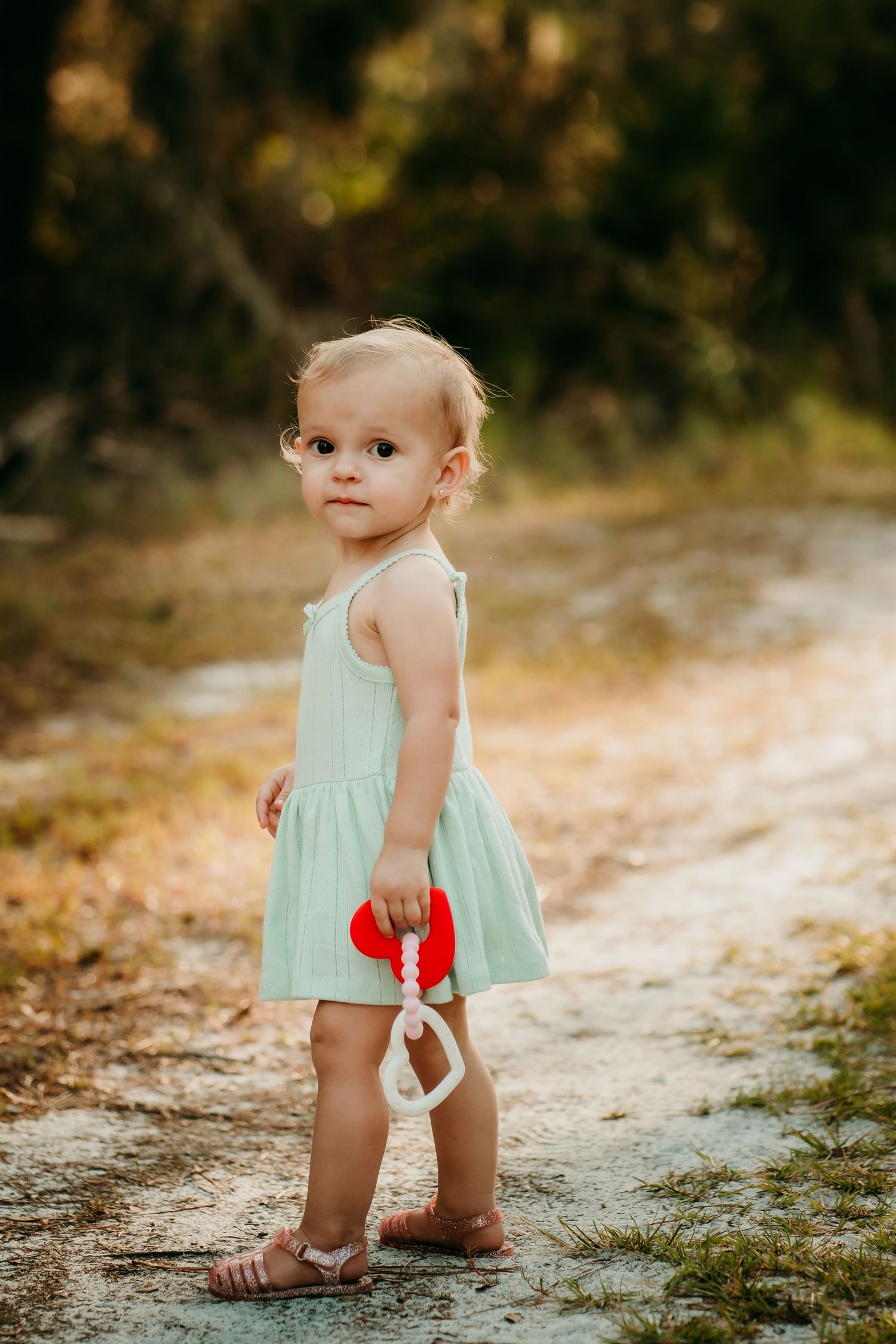 Adorable toddler girl in mint dress holding a red toy on a sandy outdoor path.