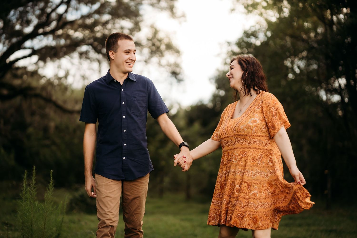 A joyful couple walks hand in hand through a lush green field, laughing together in golden hour light.
