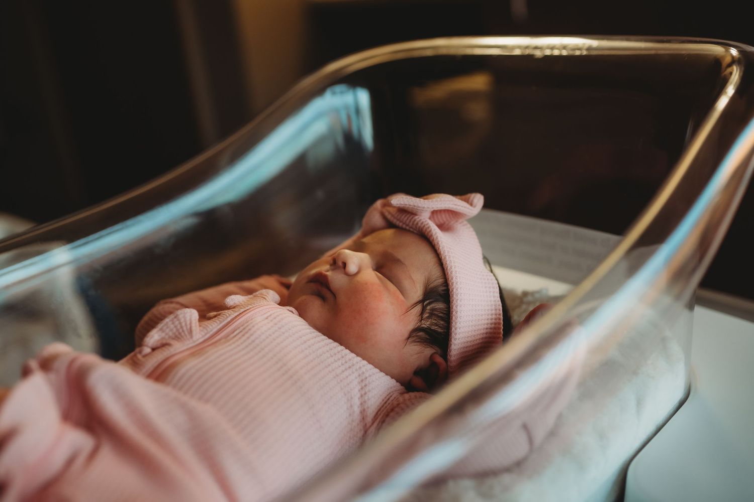 Newborn baby girl sleeping peacefully in hospital bassinet wearing pink outfit and bow headband.