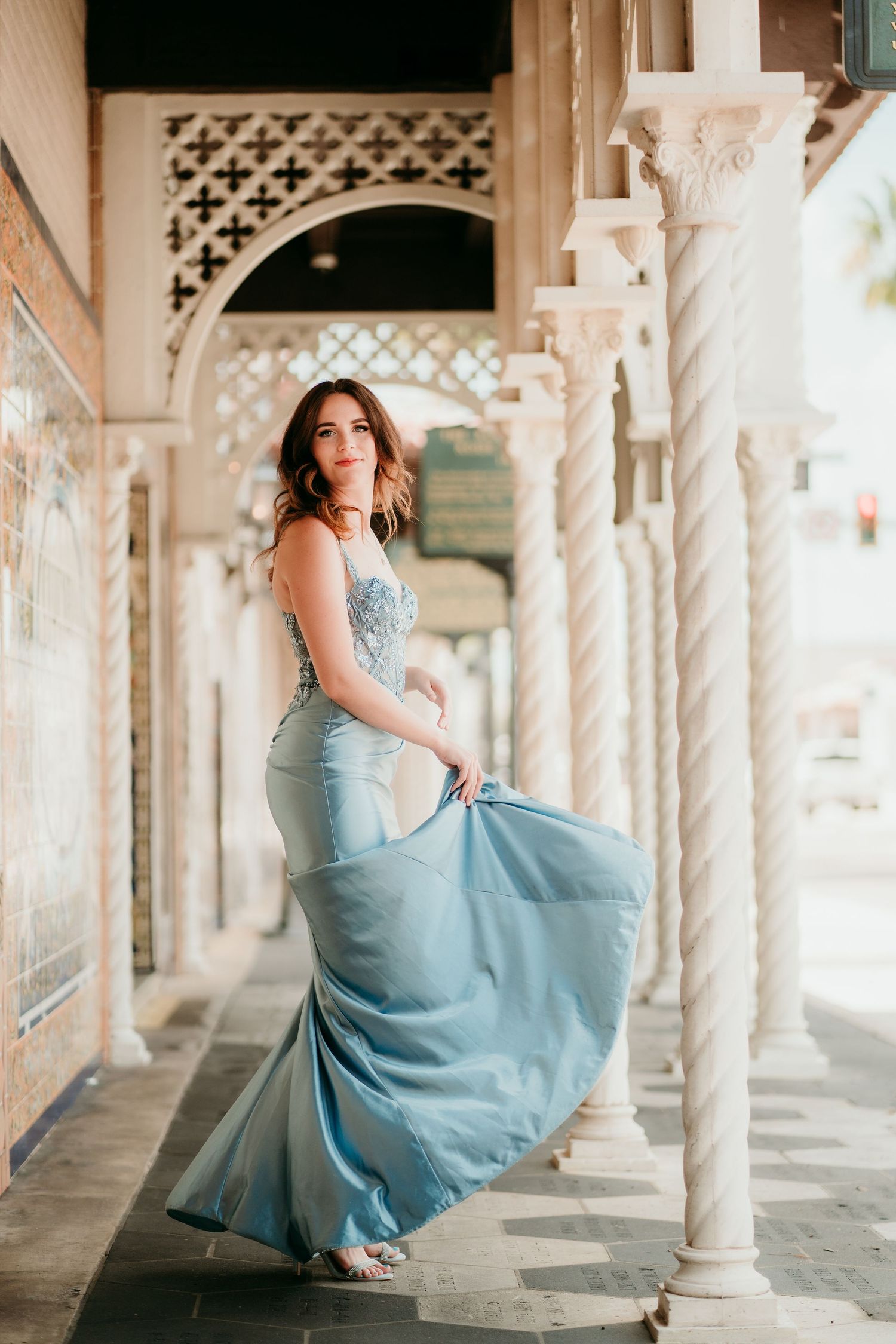 Woman in flowing blue mermaid gown posing gracefully between ornate white columns in an elegant archway.