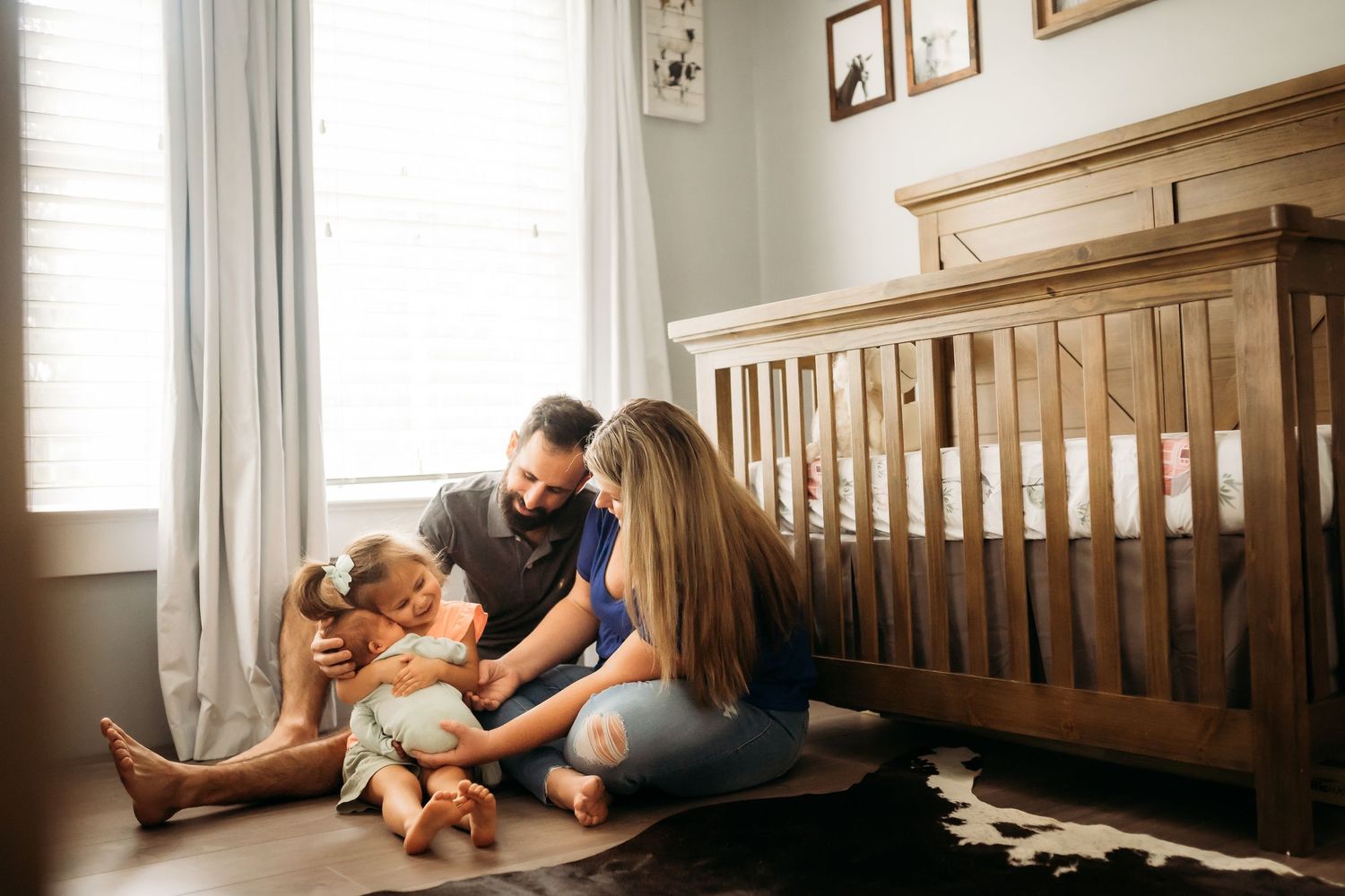 Young family sitting on nursery floor bonding with newborn and toddler by wooden crib.