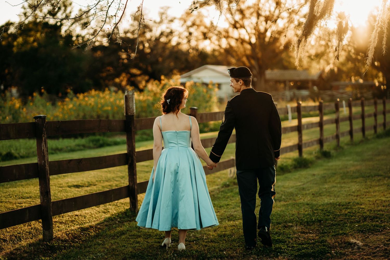 Couple walks hand in hand along wooden fence at golden hour, woman in blue dress and soldier.