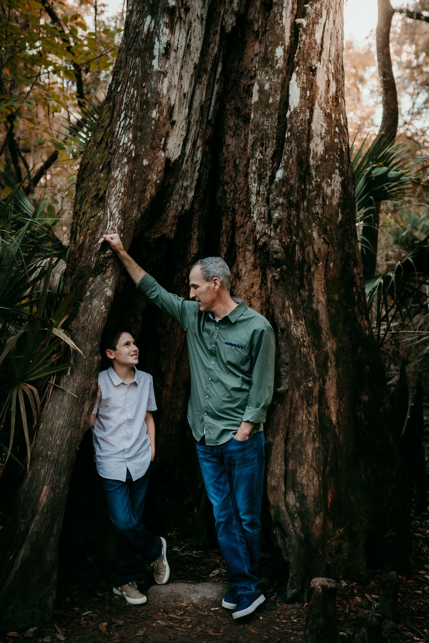 Father and son standing beneath large cypress tree in forest.