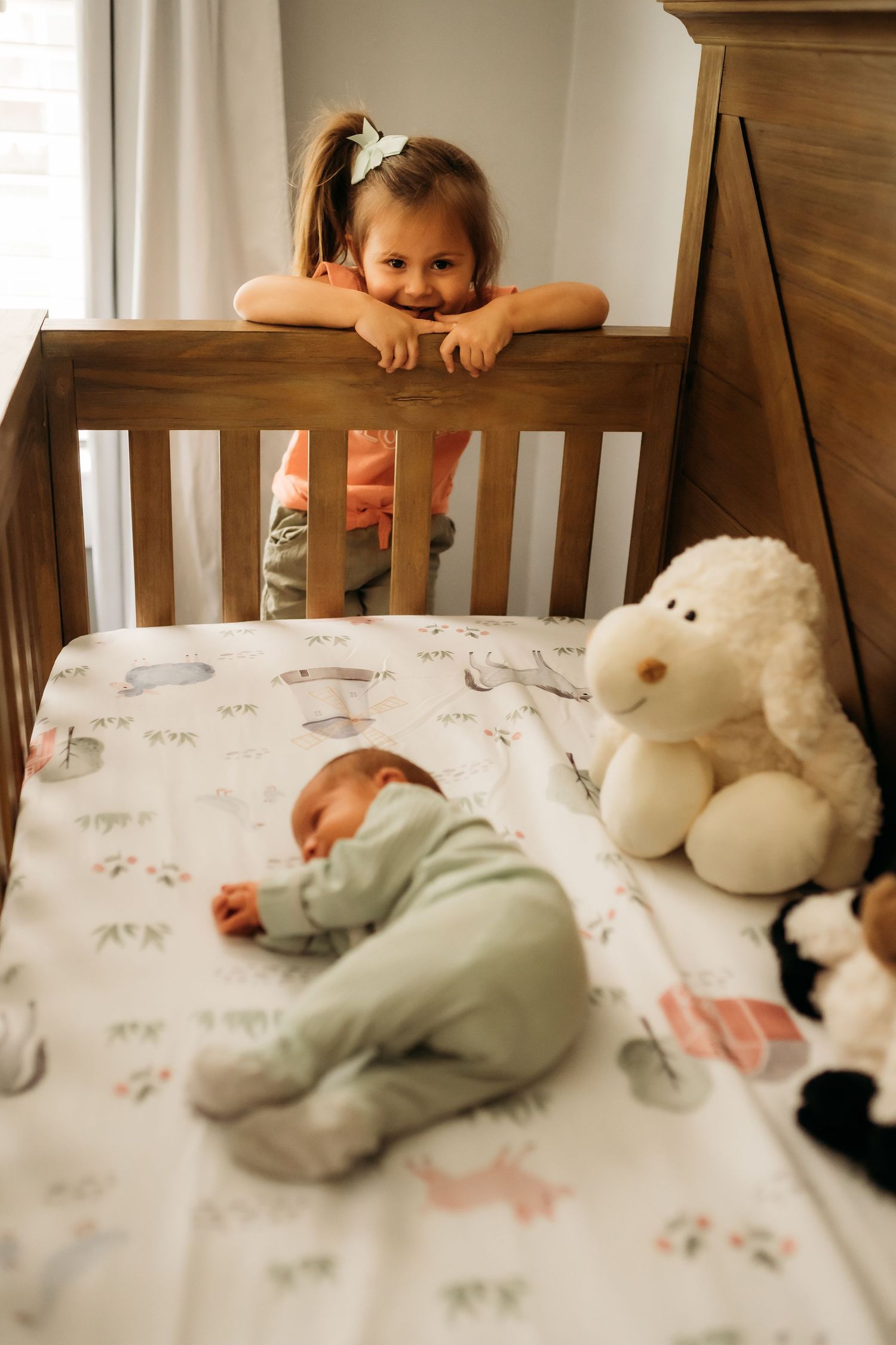 Smiling toddler girl leaning over wooden crib watching sleeping newborn baby sibling inside.