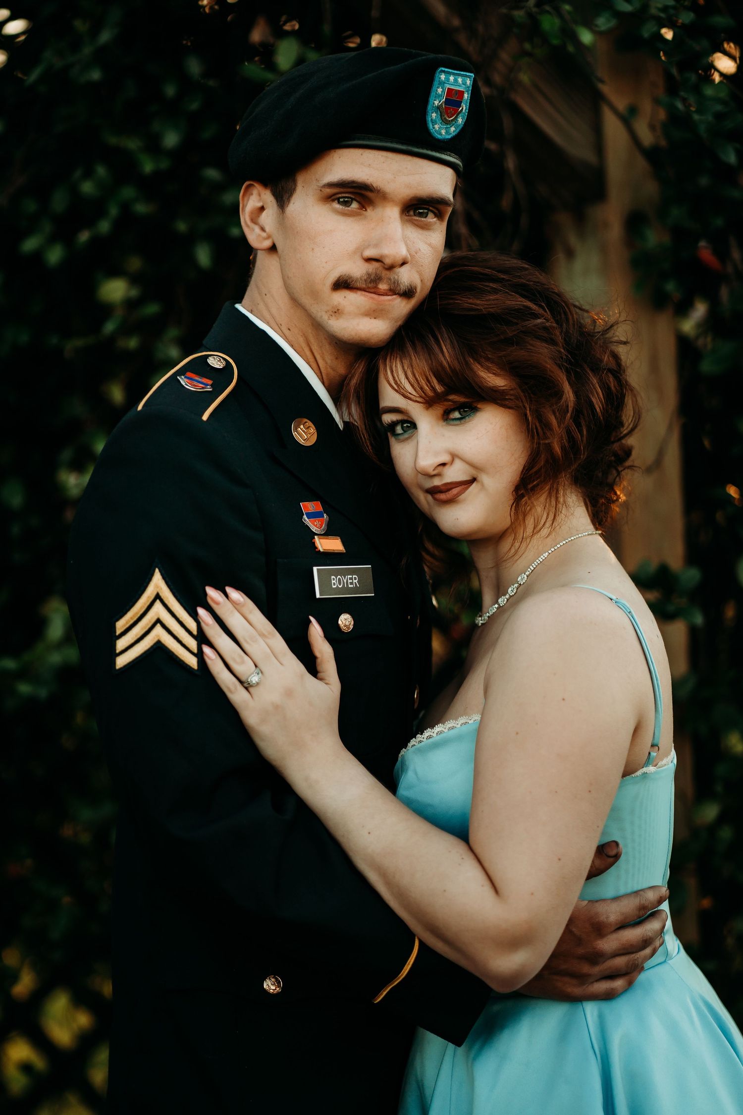 Soldier in Army dress uniform and woman in blue gown pose together in front of green foliage.